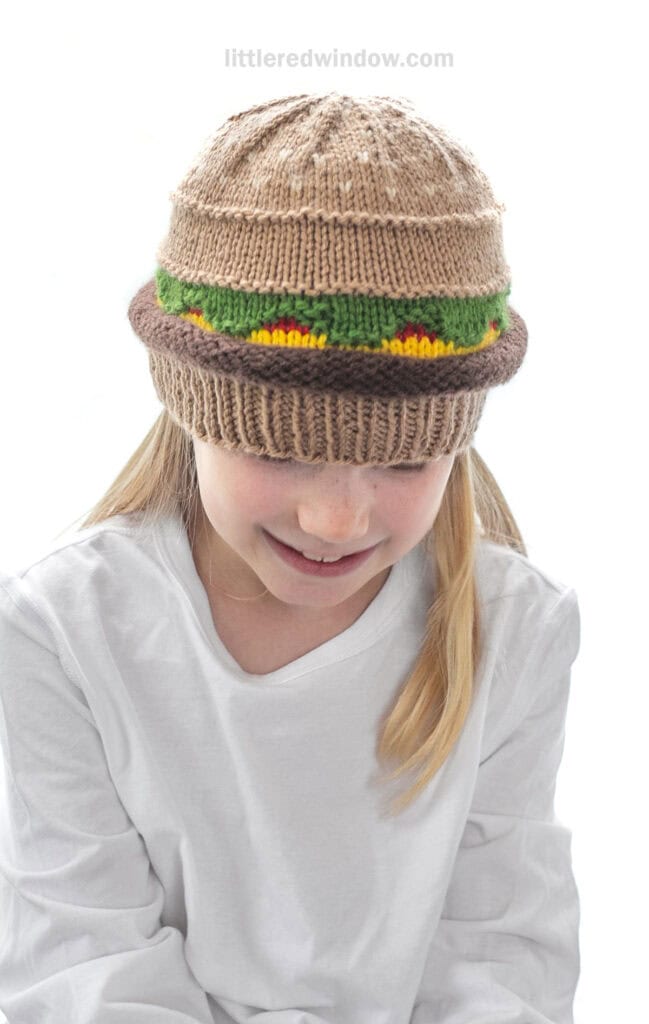 smiling girl in a white shirt wearing a knit hat that looks like a hamburger with beef, lettuce, ketchup and cheese layers looking down at their lap