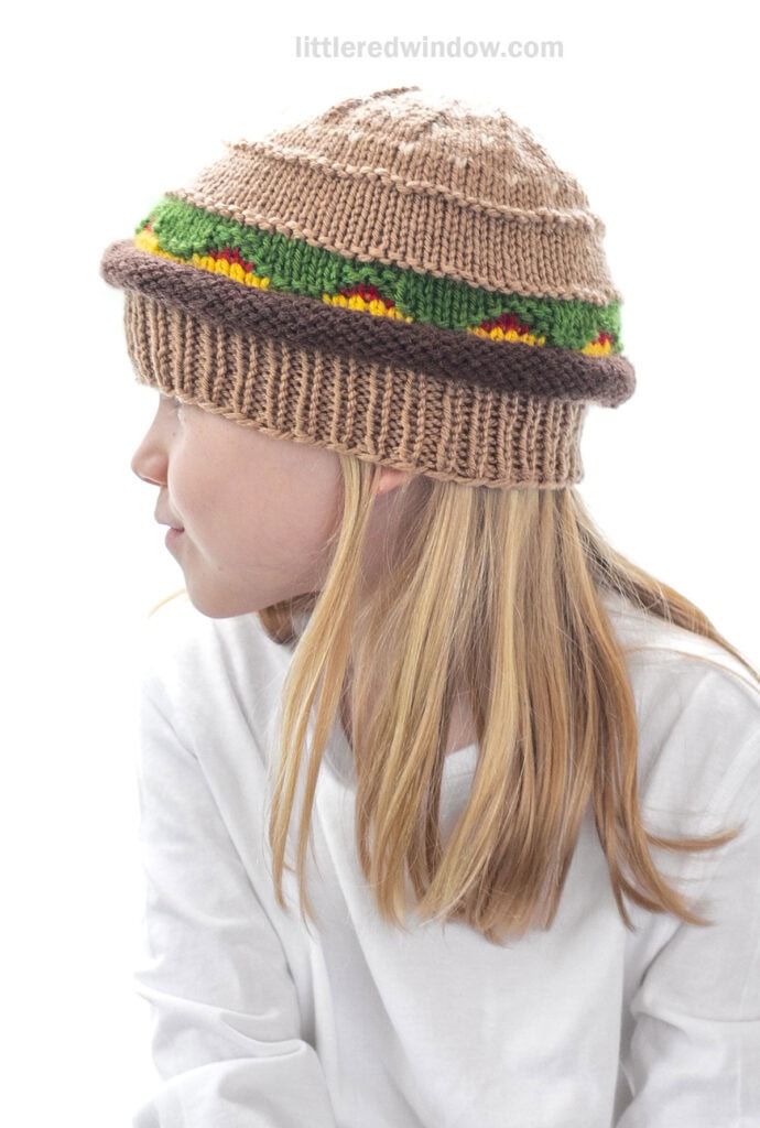 side view of child in a white shirt wearing a knit hat that looks like a hamburger with beef, lettuce, ketchup and cheese layers looking off to the left
