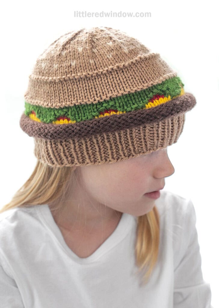 side view of child in a white shirt wearing a knit hat that looks like a hamburger with beef, lettuce, ketchup and cheese layers looking off to the right