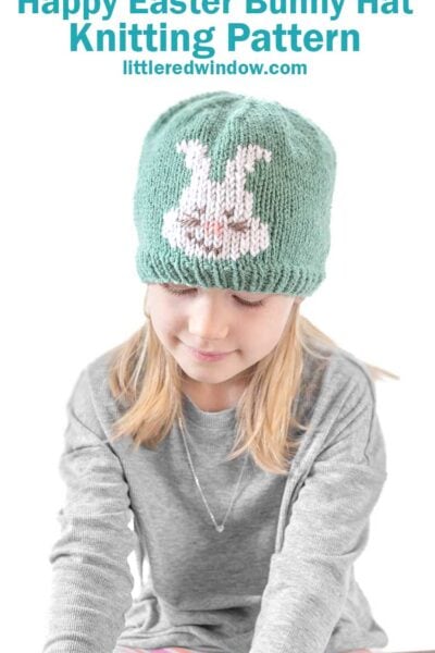 A young girl dons a green Easter bunny hat with a charming white bunny design, looking down in her cozy gray sweater. The text above says, "Happy Easter Bunny Hat Knitting Pattern littleredwindow.com.
