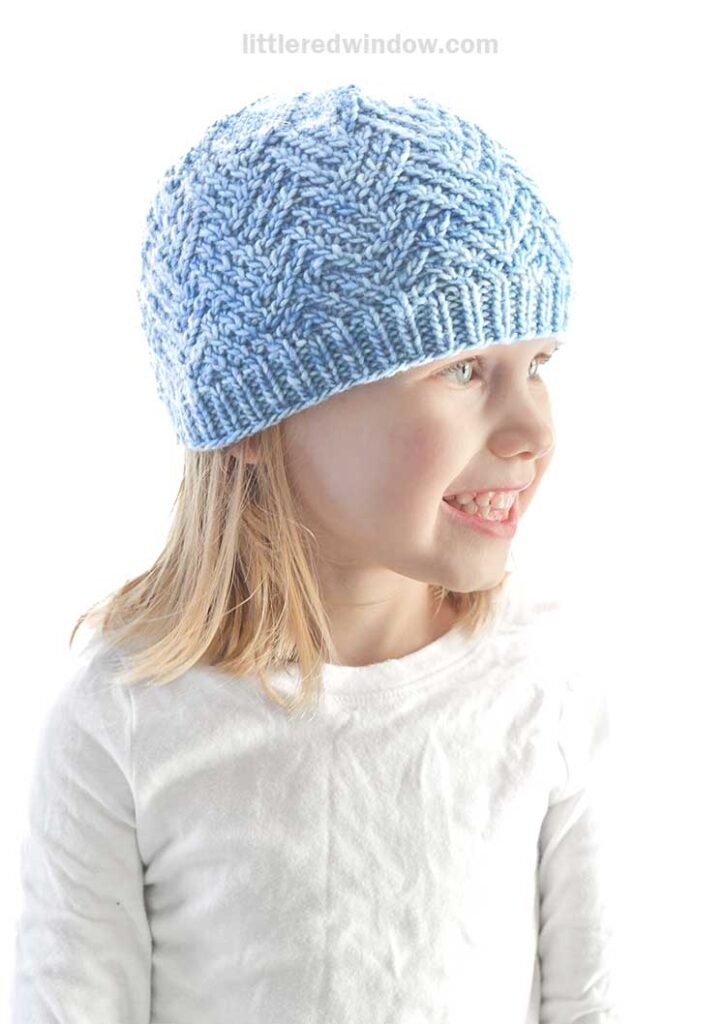 A smiling child wearing a light blue knitted hashtag hat and a white shirt. The background is bright and featureless.