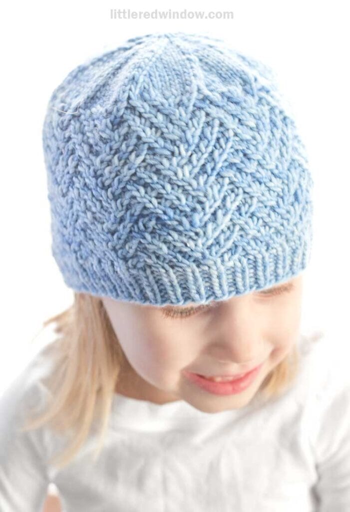 A child wearing a light blue knitted hat with a detailed pattern and hashtag smile. They're looking downwards, dressed in a plain white shirt, against a plain white background.