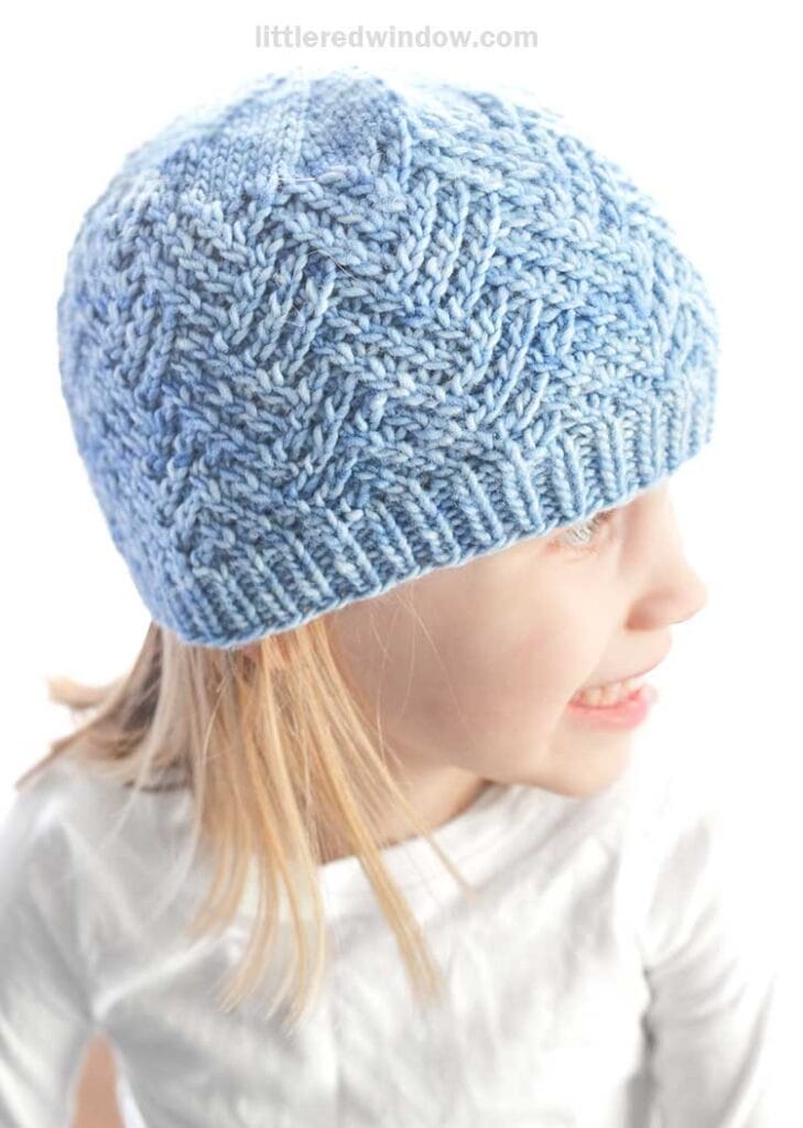 A young girl wearing a hashtag hat and a white shirt looks to the side, smiling. The light blue knitted hat features a textured, woven pattern. The softly lit background highlights her cheerful expression.