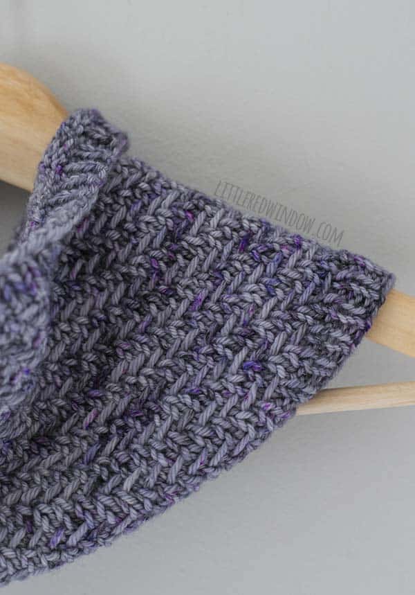 Closeup of the herringbone stitches on the hazel herringbone cowl hanging on a light wood hanger in shades of lavender and purple yarn in front of a white background