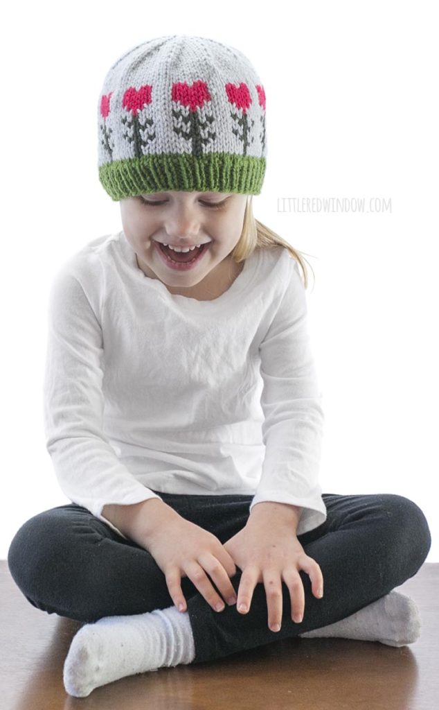 little girl in white shirt wearing light blue knit hat with pink heart shaped flowers on it looking down