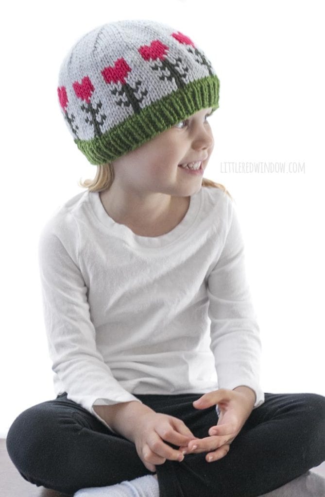 little girl in white shirt wearing light blue knit hat with pink heart shaped flowers on it looking off to the right