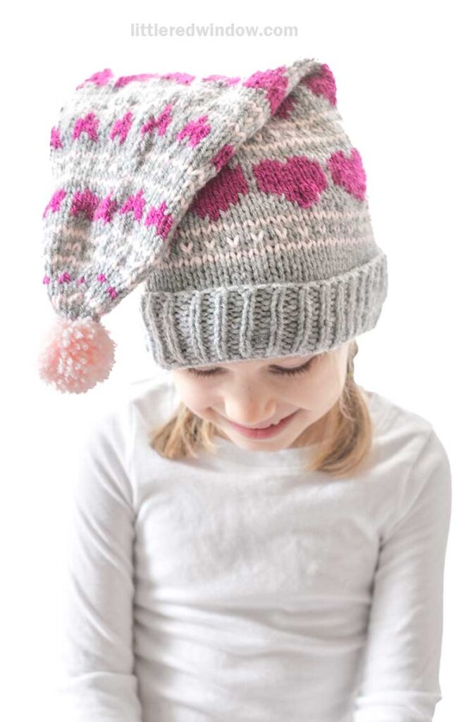A child wearing a gray knitted hat with pink heart patterns and a pom-pom, looking down with a slight smile. The child is dressed in a white long-sleeve shirt.