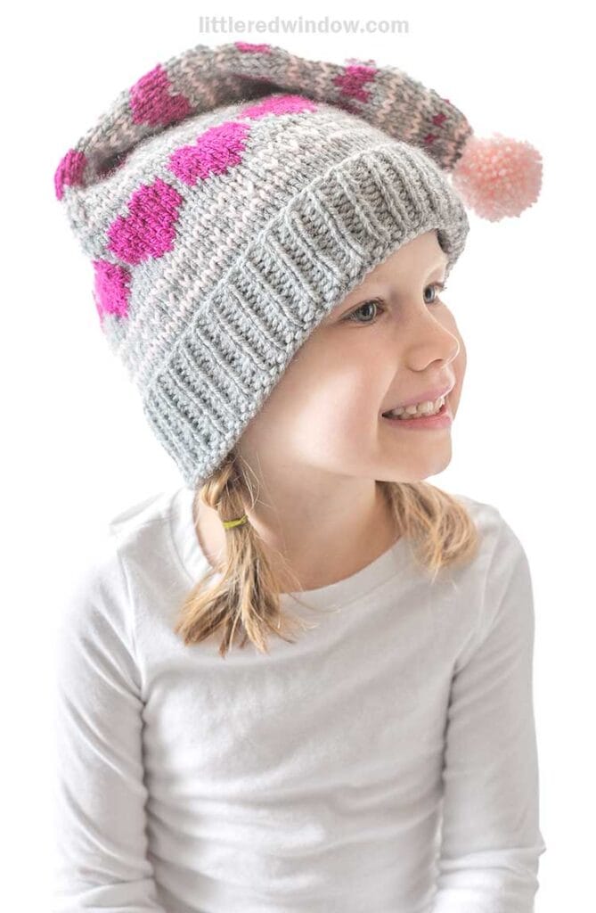 A smiling young girl wearing a knitted hat with a pattern of pink hearts and a pom-pom. She is dressed in a white long-sleeve shirt and has two small braids. The background is plain white.