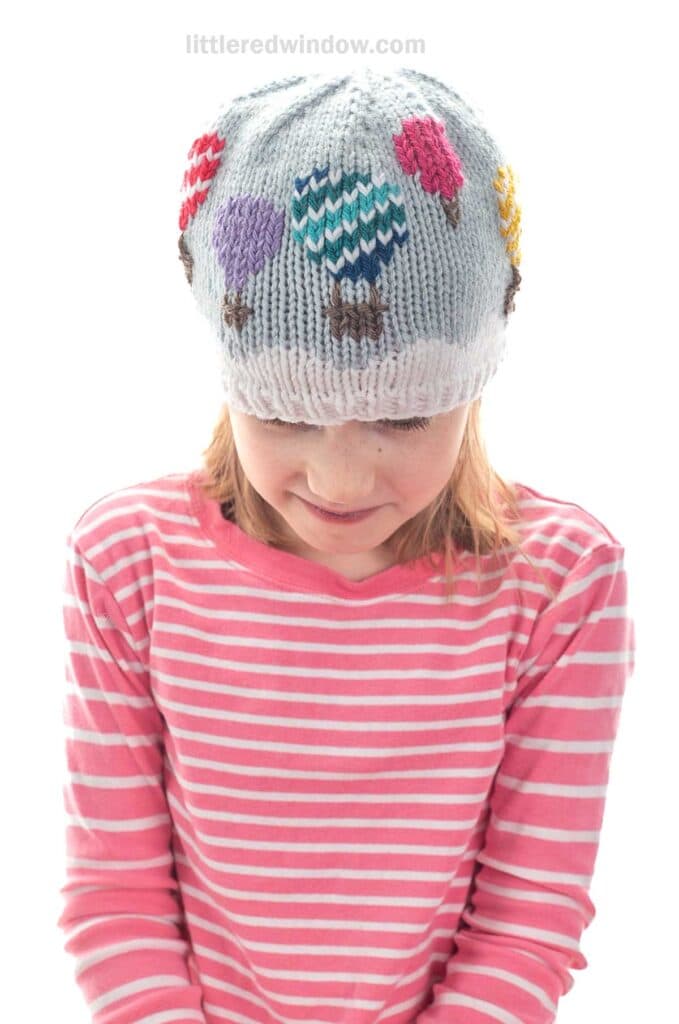 A child wearing a pink and white striped shirt and a light gray knitted hat decorated with colorful hot air balloons, looking down and smiling.