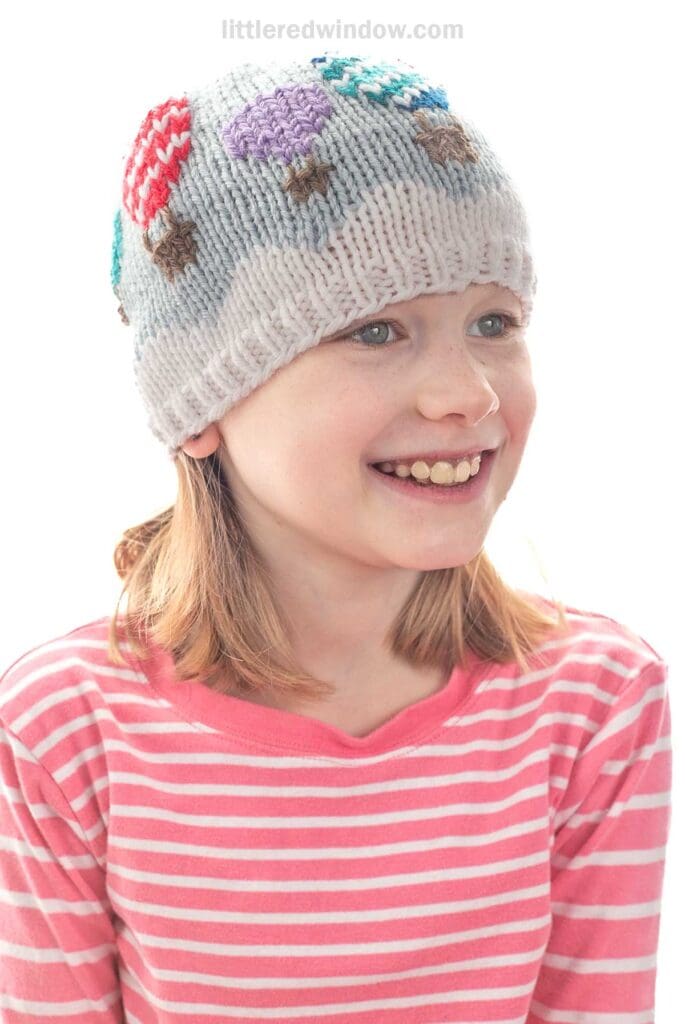 A young girl wearing a pink and white striped shirt smiles while wearing a light gray knit hat decorated with colorful hot air balloon designs.