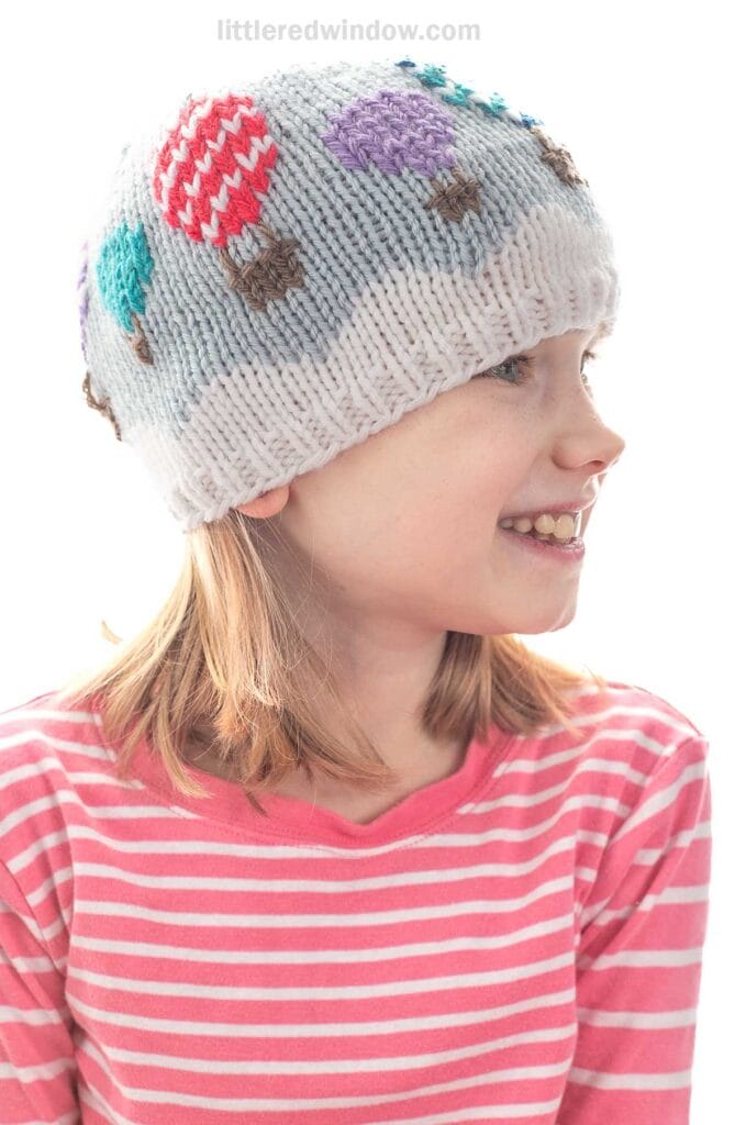 A young girl in a pink striped shirt smiles and looks to the side, wearing a light gray knit hat decorated with colorful hot air balloon designs.