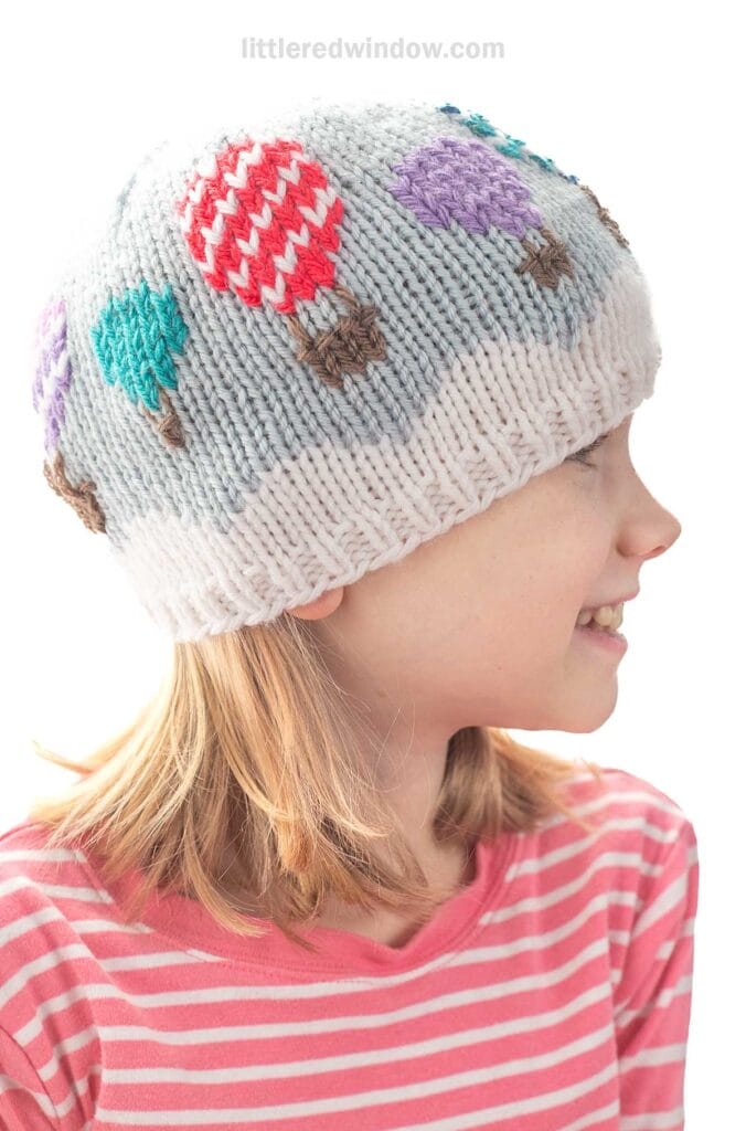 A child wearing a knit hat decorated with colorful hot air balloons looks to the side and smiles. The child has shoulder-length blonde hair and is dressed in a pink striped shirt.