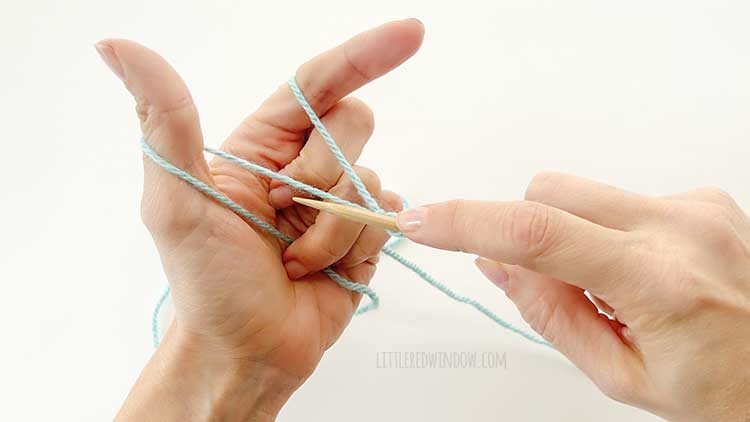 Two hands in front of a white background holding one knitting needle and light blue yarn showing german twisted cast on step 1