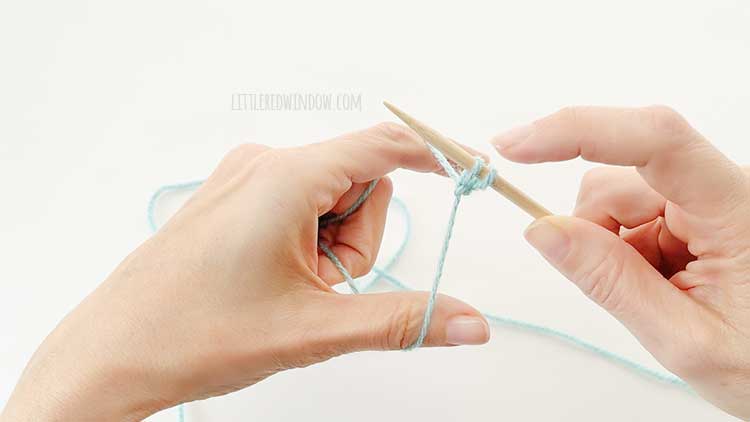 Two hands in front of a white background holding one knitting needle and light blue yarn showing german twisted cast on step 12