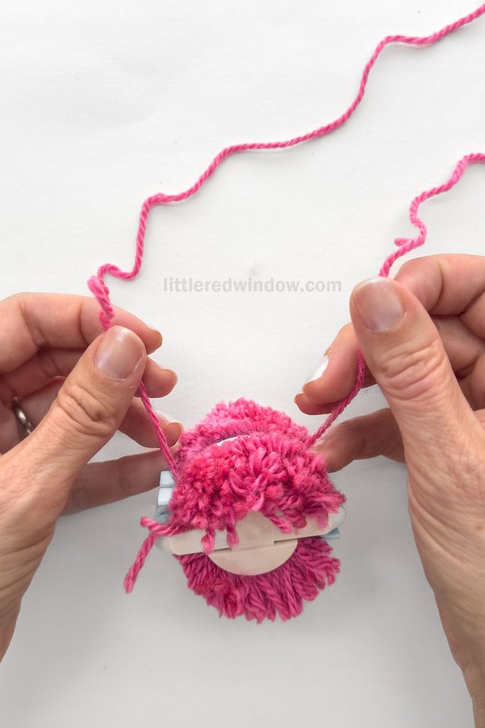 Using the piece of yarn you set aside at the beginning, wrap it around the circumference of the pom pom maker in the gap between the arches where you just cut. Pull as tightly as you can and double knot.