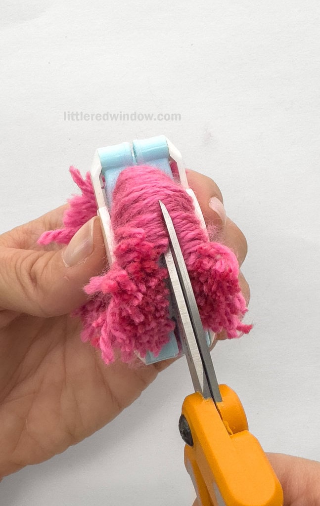Using the sharpest scissors you can find, and keeping the pom pom maker closed, cut along one edge of one half the pom pom between the gap between the arches, all the way through all of the wrapped layers of yarn.