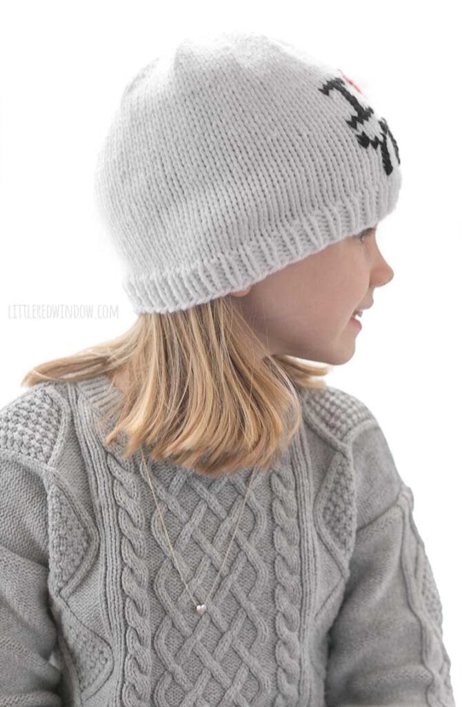 girl in gray sweater dress wearing a white knit hat with the words I heart you where the heart shape is red in front of a white background looking off to the right with a smile