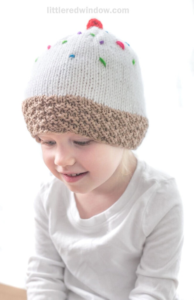 A young child is wearing a whimsical white knit hat with colorful sprinkles and a red pom-pom on top, resembling an ice cream sundae. The bottom of the hat is brown,The child is looking down and smiling, dressed in a white long-sleeve shirt.
