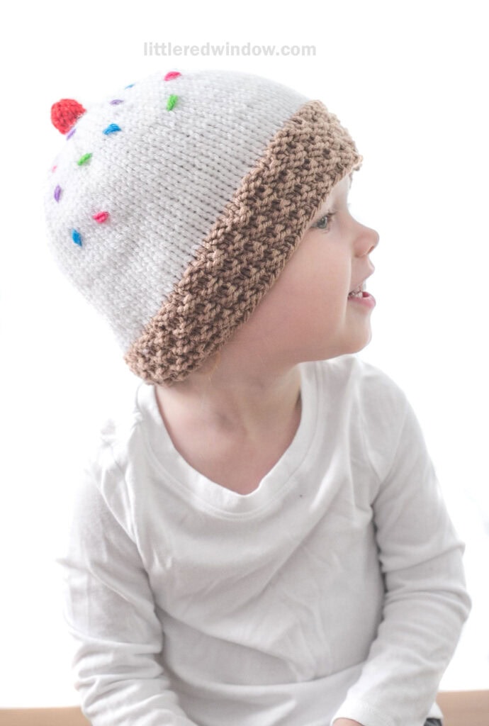 A young child wearing a knitted ice cream hat shaped like a cupcake, complete with colorful sprinkles, a red cherry on top, and a brown ribbed brim. The child is dressed in a white long-sleeve shirt and is looking to the side The background is white.