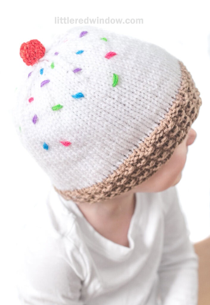 A child wearing an ice cream hat resembling a cupcake. The white hat features colorful sprinkles and a red pom-pom on top like a cherry, with a brown band at the bottom mimicking a cupcake wrapper. The child is looking slightly to the side.