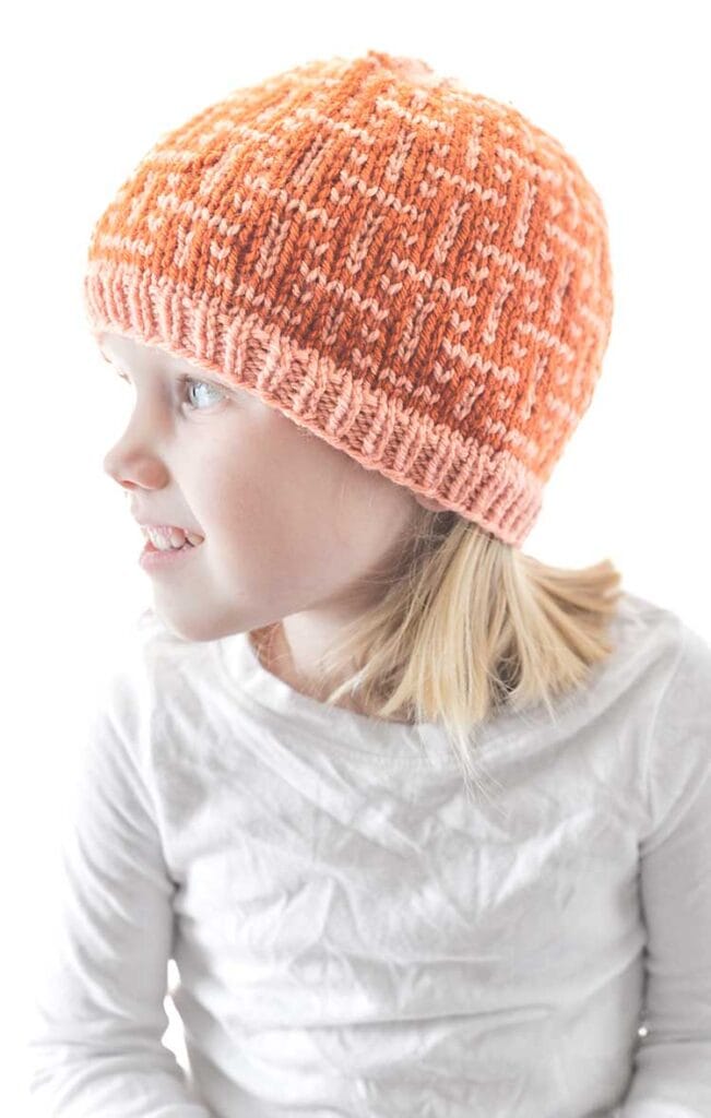 A young girl wearing an interwoven orange knit beanie and a white long-sleeve shirt looks to her left. She has shoulder-length blonde hair and is set against a plain white background.