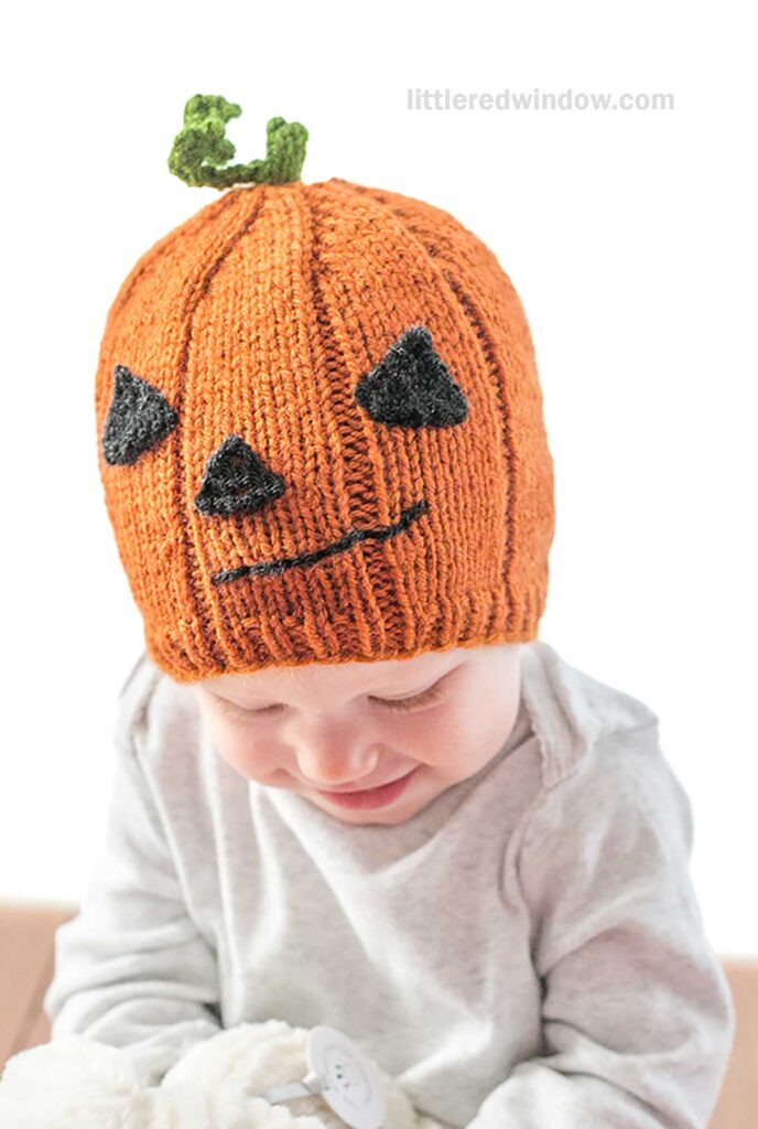 A baby wearing a pumpkin hat designed to look like a pumpkin, complete with a green stem on top and black triangular eyes and a crooked mouth. The baby, dressed in a light gray outfit, is looking down while holding a stuffed animal.