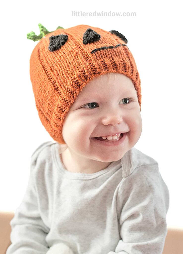 A smiling baby wearing a light-colored long-sleeve shirt and an orange pumpkin hat designed to look like a jack-o'-lantern, complete with a green stem on top, sits against a white background. The hat features black eyes and a mouth.