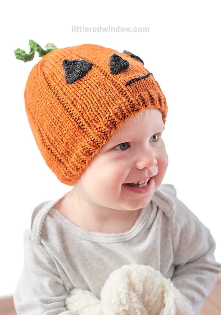 A smiling baby wearing a pumpkin-themed knit hat with a green stem on top and a black jack-o'-lantern face. The baby, dressed in a light gray outfit, is holding a soft, cream-colored toy or blanket. The adorable pumpkin hat adds a festive touch, while the background remains plain white.