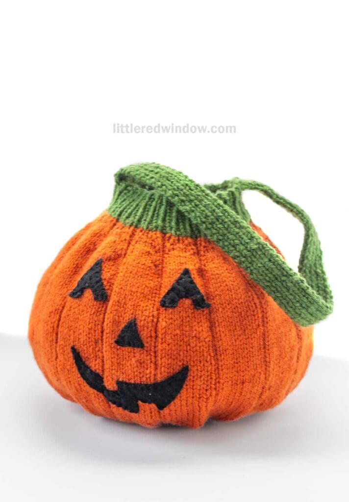 A knitted orange Jack-o'-Lantern Trick-or-Treat Bag with a green handle and a black jack-o’-lantern face, displayed against a white background.