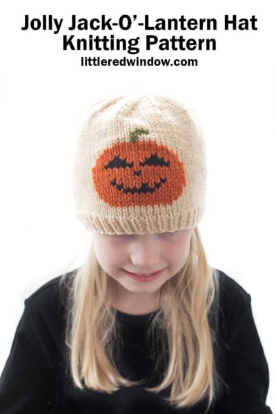 Blond girl wearing a black shirt and a tan knit hat with a smiling jack o lantern in duplicate stitch on the front center while she is smiling and looking down in front of a white background