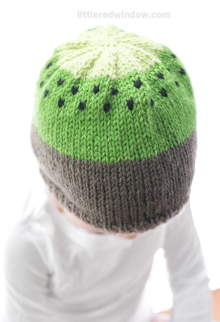 little girl wearing a knit hat that looks like a kiwi fruit and looking down