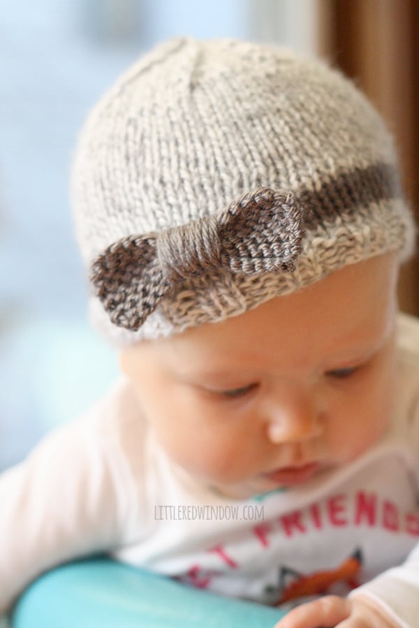 baby looking down wearing gray knit hat with purple stripe and purple knit bow