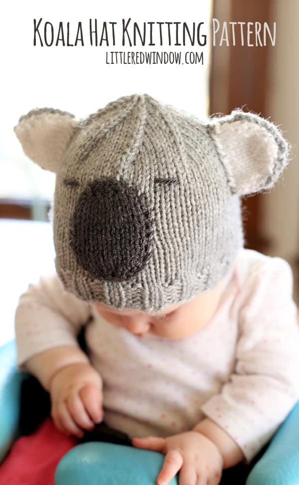 Baby in a pink and white shirt sitting in a blue chair wearing a gray knit hat that looks like a koala looking down and to the left
