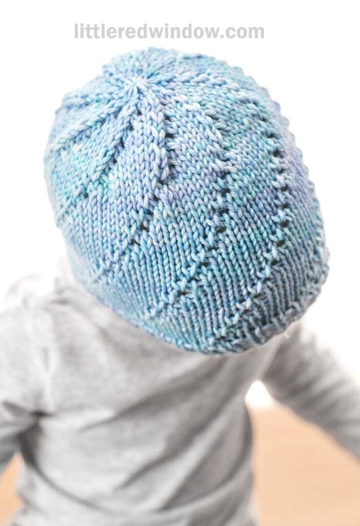 A child wearing a light blue, hand-knitted Lace Twist Hat with intricate patterns. The child is facing away from the camera, showcasing the top and back of the hat. The background is blurred, emphasizing the hat's detail. The website name