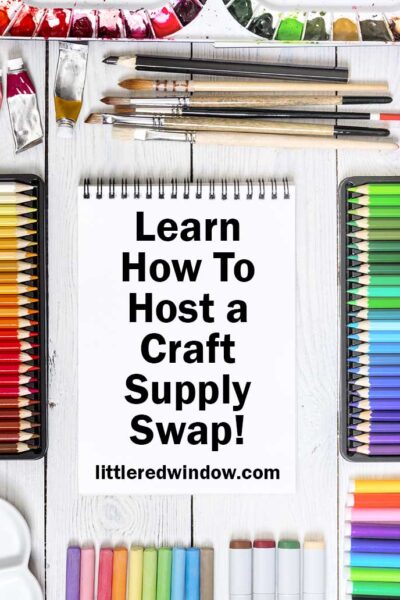 lines of rainbow colored pencils and paintbrush arranged around a white paper notebook on a white wood tabletop