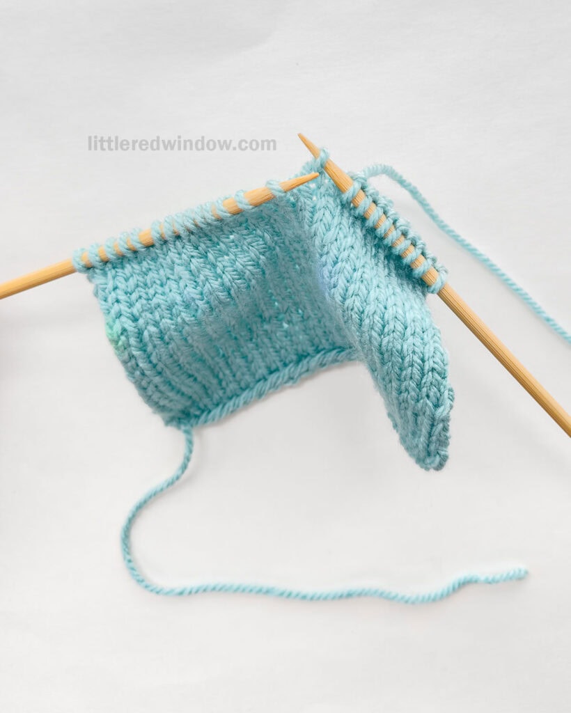 two wood knitting needles and a blue knit stockinette swatch in front of a white background