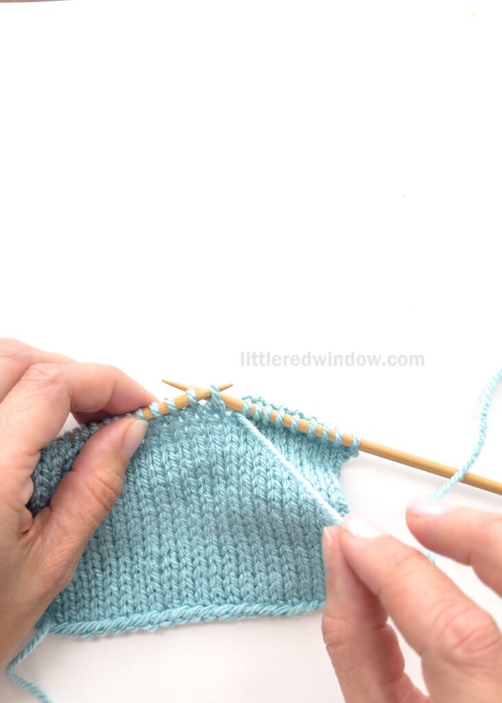 hands holding a swatch of blue knitting on wood knitting needles with the right hand holding the working yarn IN FRONT of the work