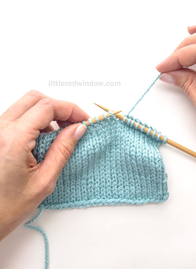 hands holding a swatch of blue knitting on wood knitting needles with the right hand holding the working yarn IN BACK of the work