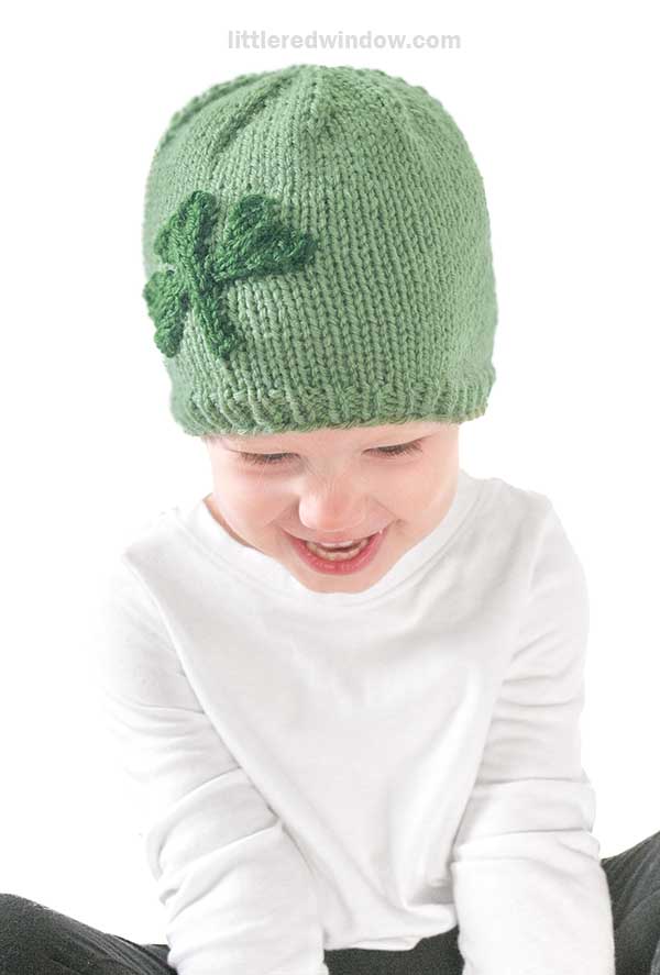A child wearing a shamrock hat smiles while looking down, dressed in a white long-sleeve shirt against a plain white background.