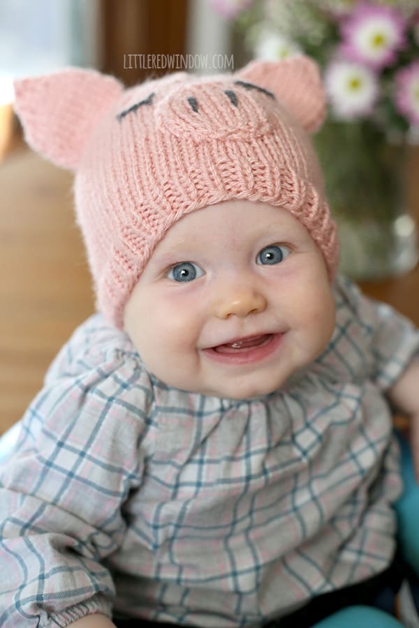 Smiling baby in plaid shirt wearing a pink knit pig hat looking up at the camera