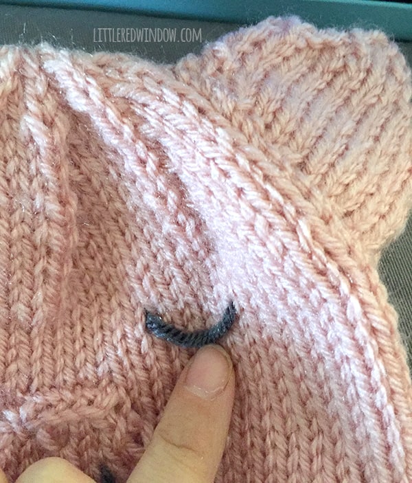 Hand pointing at how to embroider the eye on a pink knit pig hat