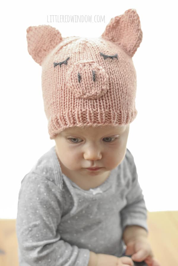 Toddler in a gray shirt wearing a pink knit pig hat and looking over their right shoulder