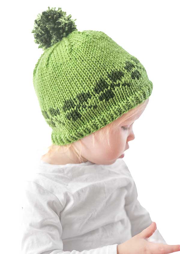 A young child wearing a charming shamrock hat featuring a knitted green pom-pom and dark pattern near the brim. The child is dressed in a crisp white shirt against a plain background, as they gaze downward in quiet contemplation.