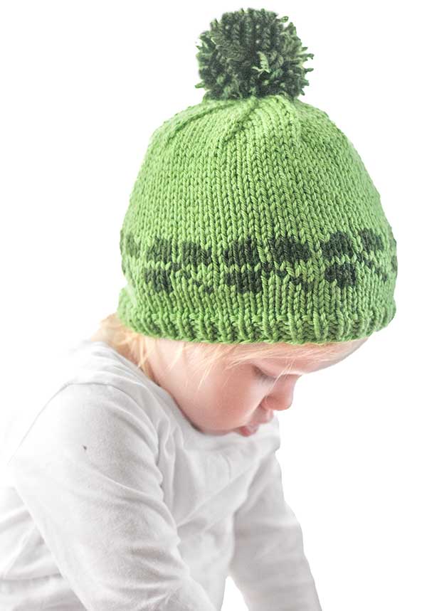A toddler wearing a whimsical shamrock hat, adorned with a pom-pom and darker green dots, gazes downward. The child's white top contrasts against the plain white backdrop.