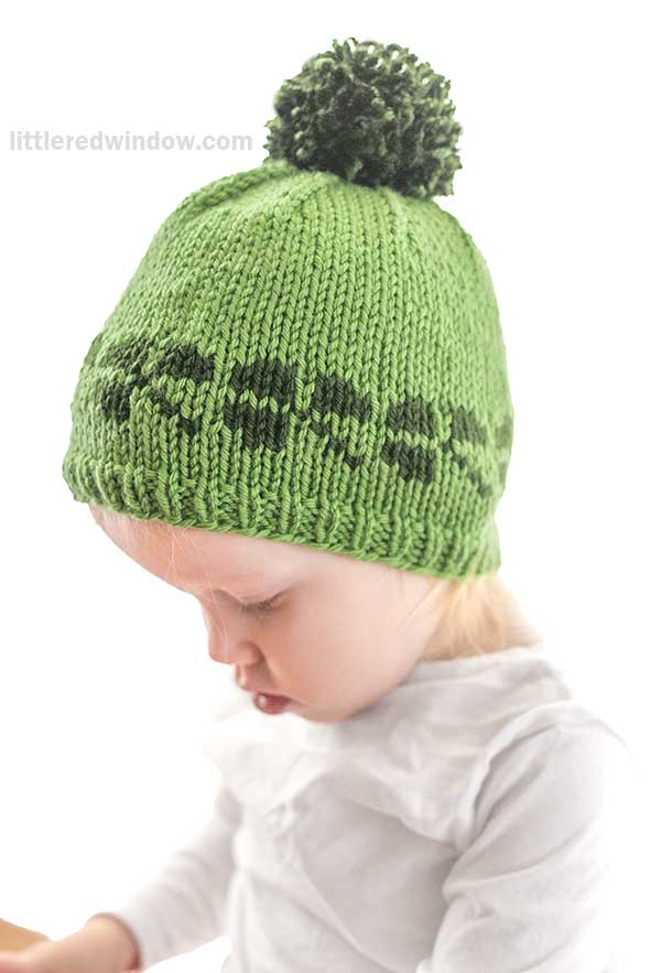 A toddler wearing a green knit shamrock hat with a pom-pom and a dark green pattern near the brim, looking downward. The background is white, and the child is dressed in a white shirt.