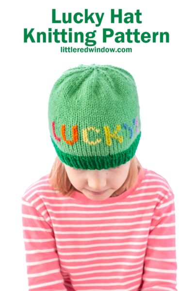 Child wearing a green knitted hat with the word "LUCKY" stitched in colorful rainbow letters. The child is dressed in a pink and white striped shirt. Text above reads "Lucky Hat Knitting Pattern littleredwindow.com.