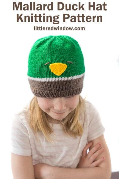 girl wearing a knit hat in brown white and green that looks like a mallard duck looking down at her lap
