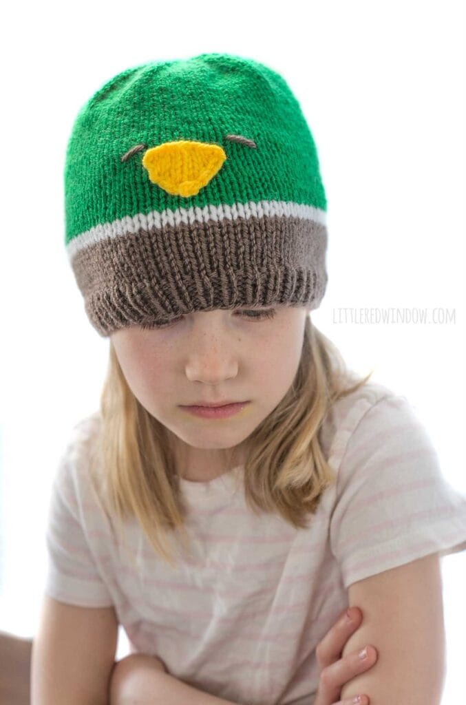 girl wearing a knit hat in brown white and green that looks like a mallard duck looking down at her lap