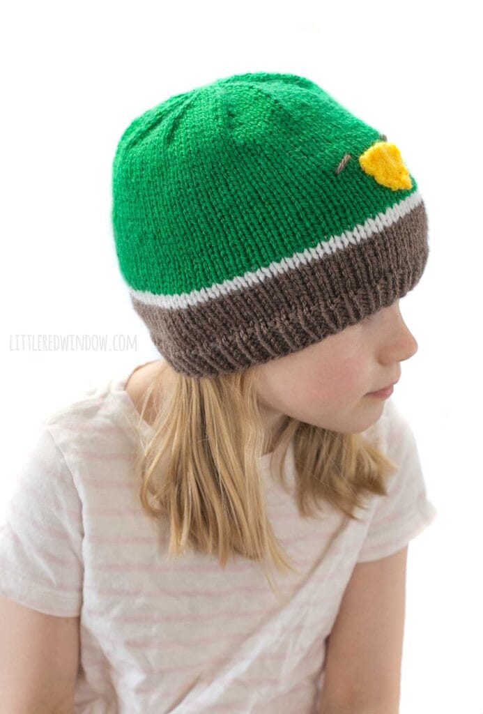 girl wearing a knit hat in brown white and green that looks like a mallard duck looking off to the right
