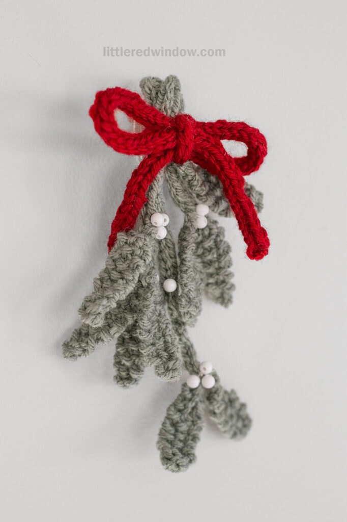 A crocheted green mistletoe with white berries, crafted using a mistletoe knitting pattern, is tied with a red yarn bow and hangs against a plain white background.